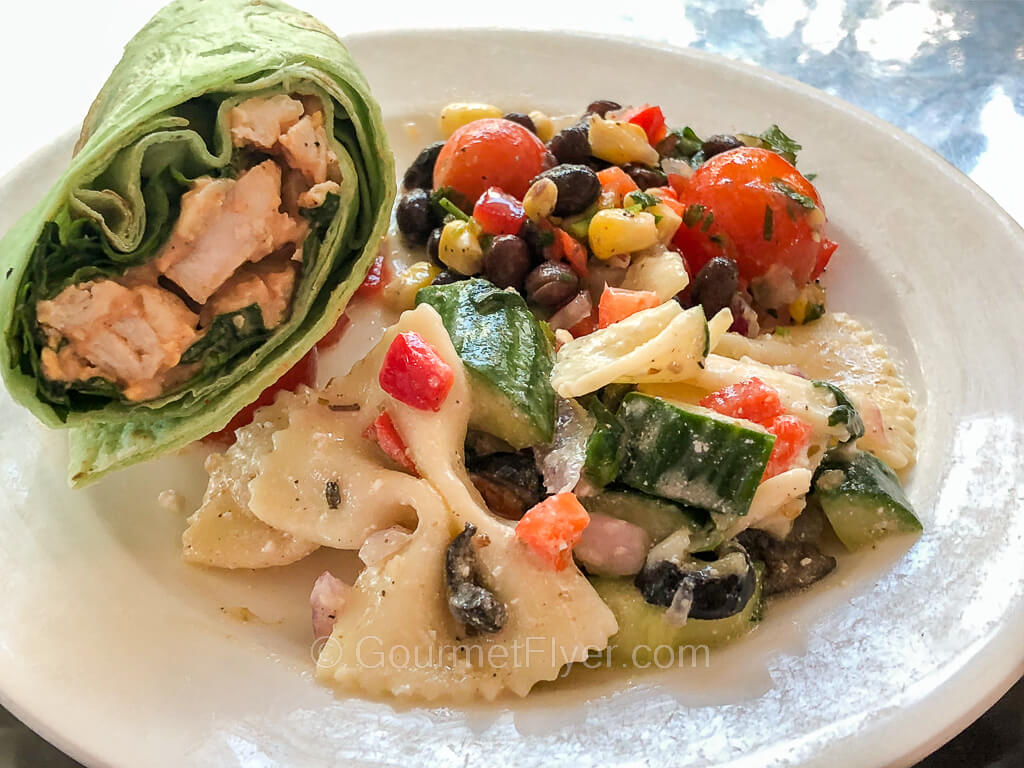 A chicken roll in a green veggie wrap is served on a plate along with pasta salad and corn and black beans.