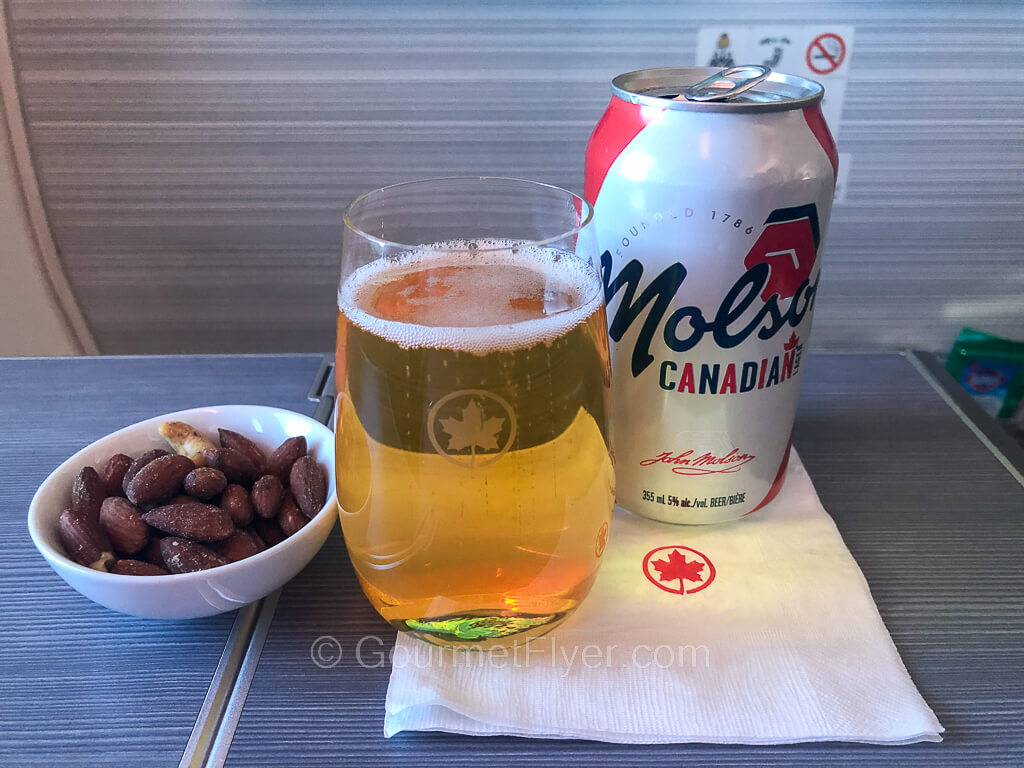 A can of Molson Canadian is accompanied by a full glass of beer and a small dish of nuts.