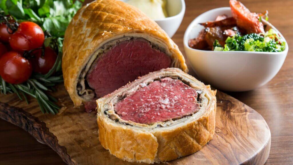 A thick slice of beef Wellington is cut from a roll and served on a cutting board.