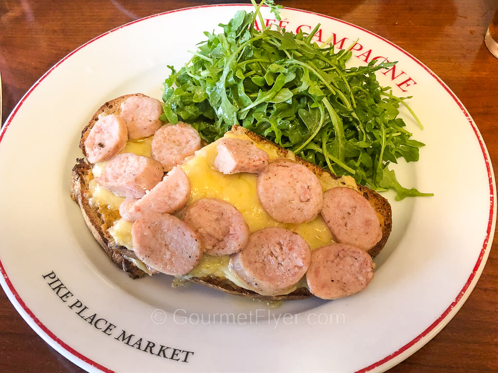 An open-faced sandwich is topped with sliced sausage and melted cheese and accompanied with arugula salad.