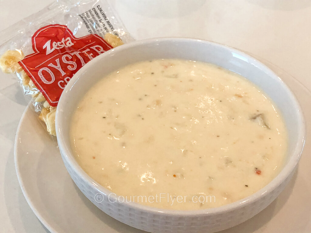 A bowl of clam chowder is served on a plate with a packet of oyster crackers.