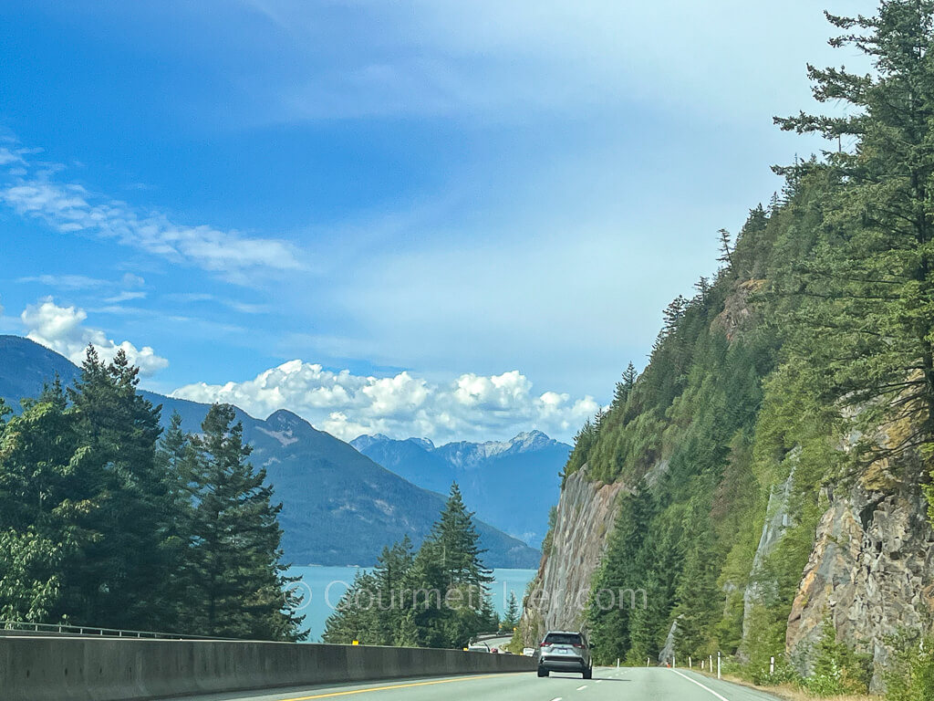 A two-lane divided highway has views of sea and mountains on the left-hand side on a mostly sunny day with few clouds.
