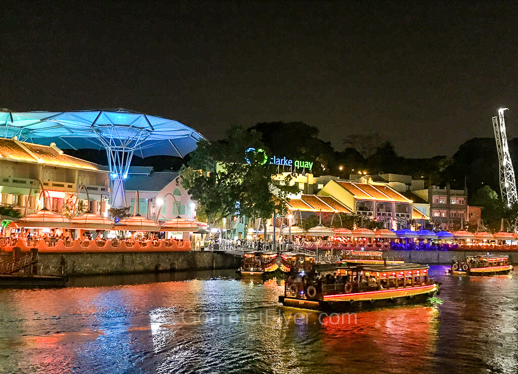 Boats filled with tourists cruise along the Singapore River with the colorful lightings of Clarke Quay in the background.