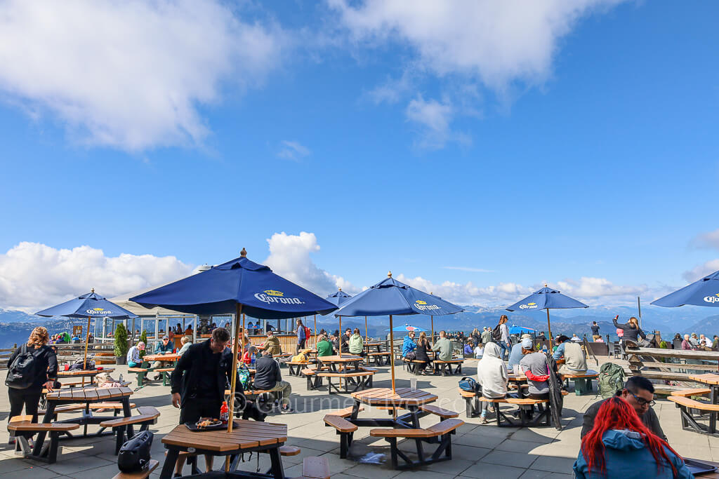 An outdoor dining area with tables covered by umbrellas on a sunny day with blue sky and few clouds.