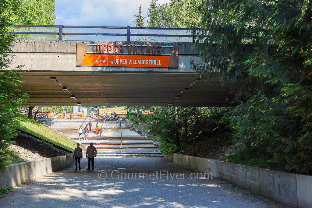 The "Upper Village" sign hangs on an overpass on top of a path leading to a set of wide stairs.