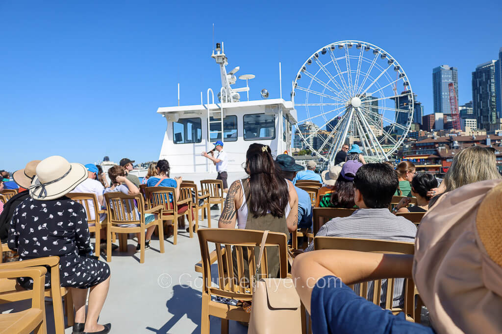 Review of the Seattle Harbor Cruise features engaging tourists on the open-air sun deck as the boat turns to face the Ferris wheel.