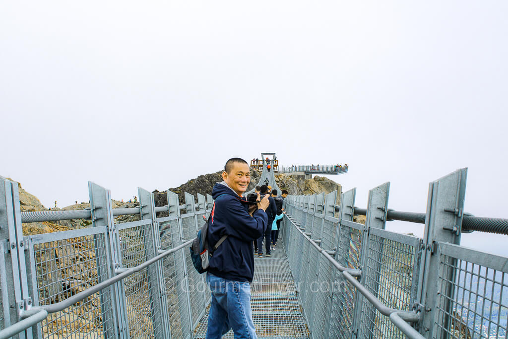 The Gourmet Flyer turns around while crossing the suspension bridge, holding a camera in his hands.