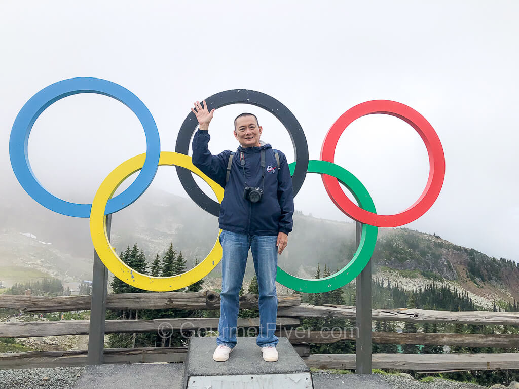 The Gourmet Flyer is standing on the podium with the 5 colorful Olympics rings, waving his right hand.