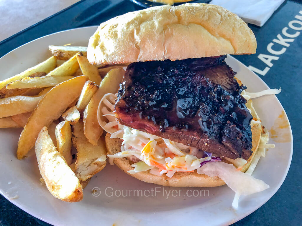 A BBQ tri tip sandwich is served on top of a bed of coleslaw in a bun and accompanied by French fries on a plate.