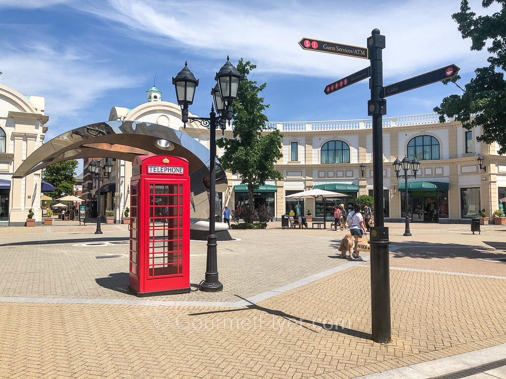 A British style red phone booth is installed in a trendy open space in an outdoor mall, with a large two-story building in the background.