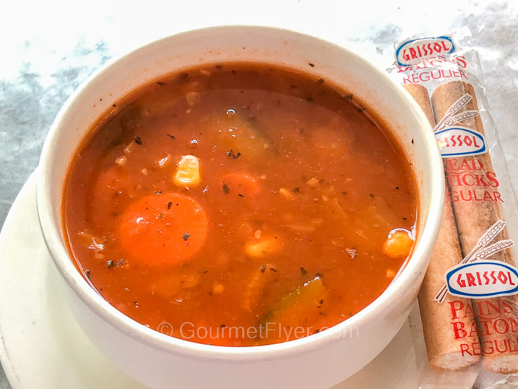 A bowl of minestrone soup sits on a plate with a package of two mini breadsticks.