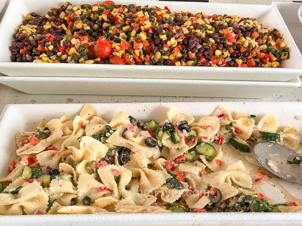 In the front is a long serving platter of pasta salad, and in the back, a black beans and corn salad.