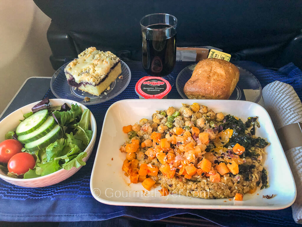 A dinner tray contains a dish of quinoa topped with butternut squash, a side salad with sliced cucumber and cherry tomatoes, dessert, a roll, and a glass of red wine.
