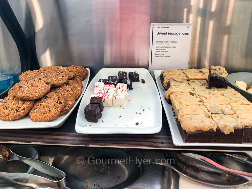 Three long trays of desserts are served on a counter. From left, cookies, petite white and dark chocolate cakes, and squares of a chocolate sponge cake.