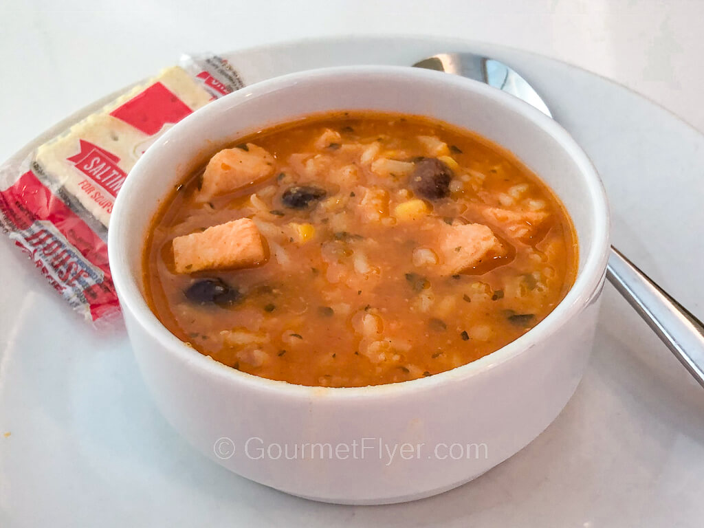 A bowl of tomato-based soup is loaded with chunks of chicken meat, rice, and black beans. A packet of crackers is served on the side.