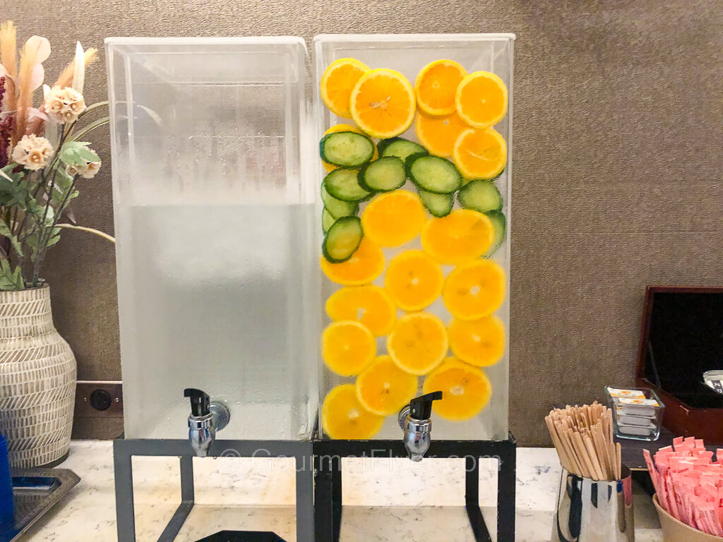 Two tubs of water sit atop a counter; one of them has slices of oranges and cucumbers in it.