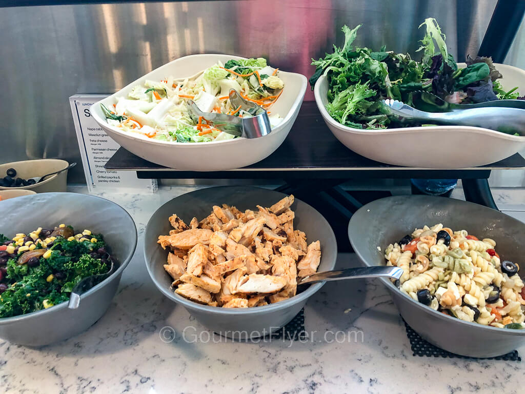 Two serving bowls of greens are on the top shelf of the counter. The lower shelf has bowls of greens, chicken meat, and pasta salad.