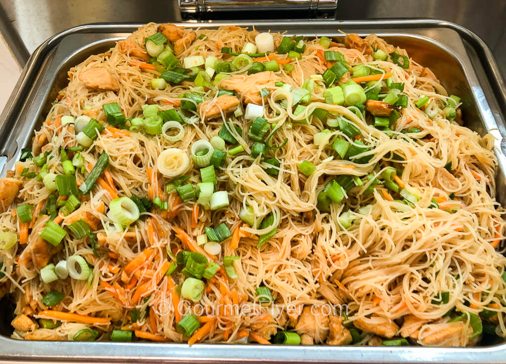 A full serving pan of fried thin rice noodles sauteed with chicken strips is sprinkled with chopped green onions.