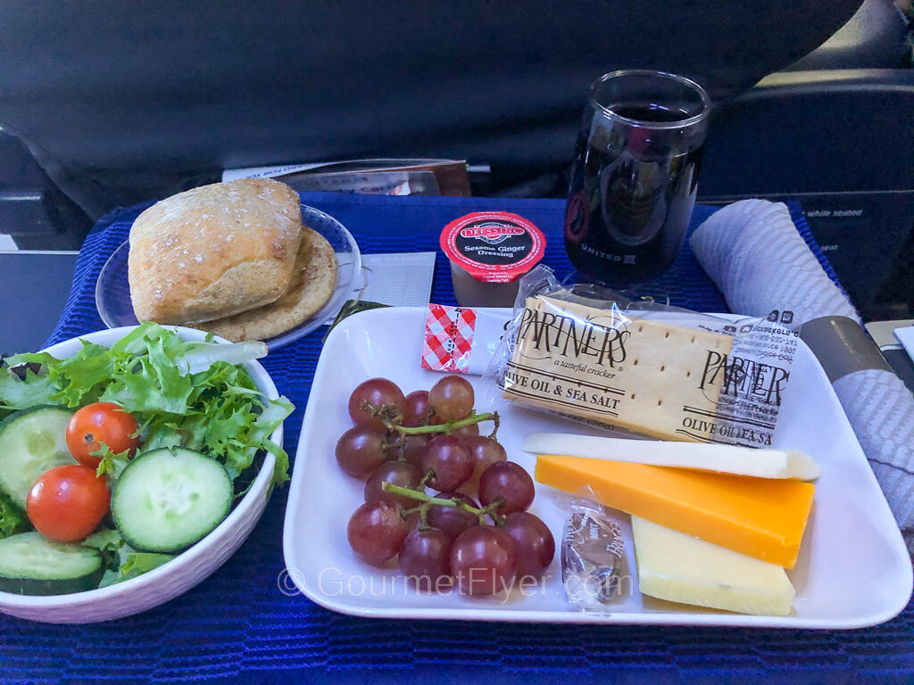 A dinner tray has a plate of cheeses, crackers, and grapes, a green salad, and a dinner roll. It is accompanied by a glass of red wine.