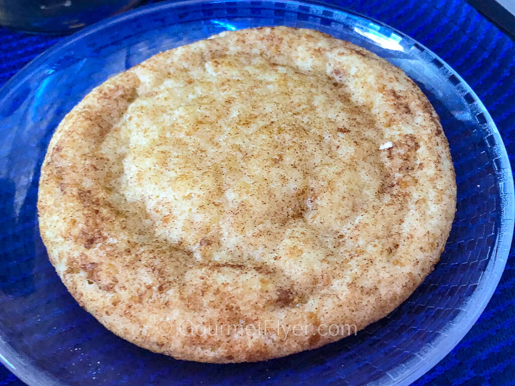 A white chocolate cookie sits on a plate atop a dinner tray with blue tablecloth.