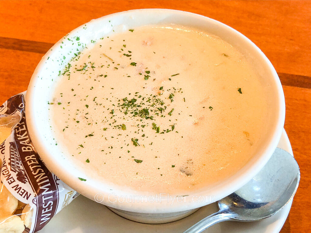 A small bowl of creamy chowder is sprinkled with parsley on top and served with a packet of oyster crackers.