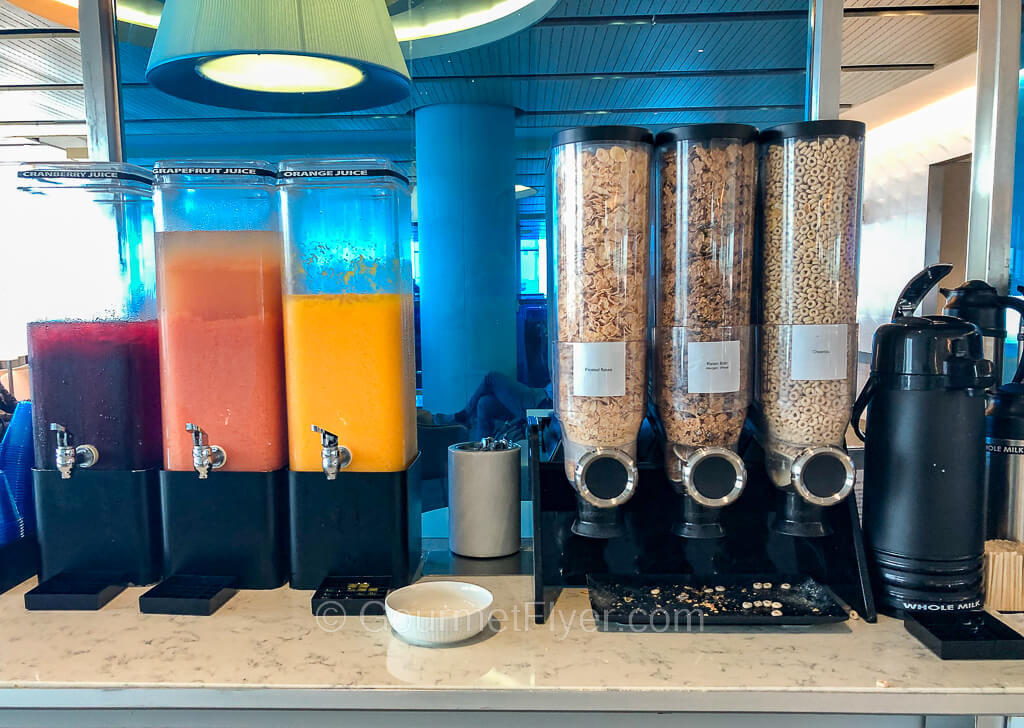 Three tall glass canisters to the left are filled with cranberry, grapefruit, and orange juices, while three other ones to the right are filled with cereals.