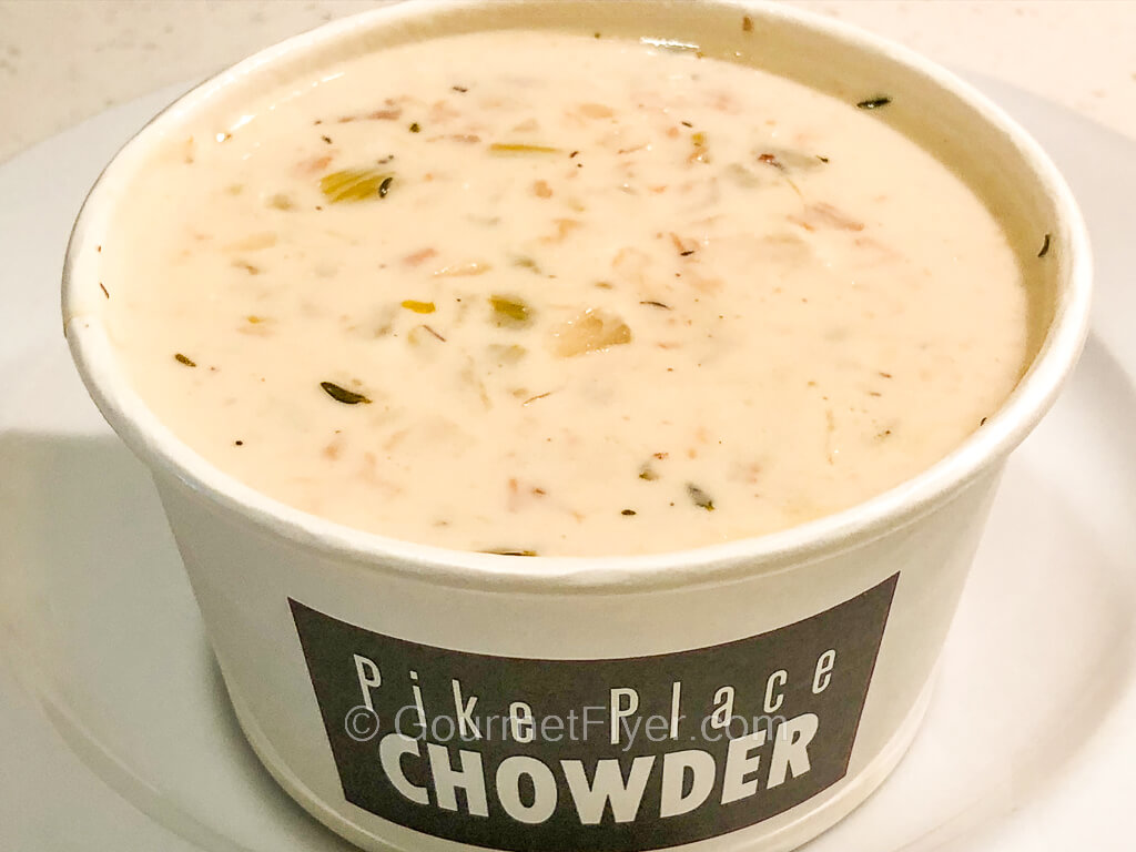 A creamy chowder is served in a paper bowl with the logo of Pike Place Chowder.