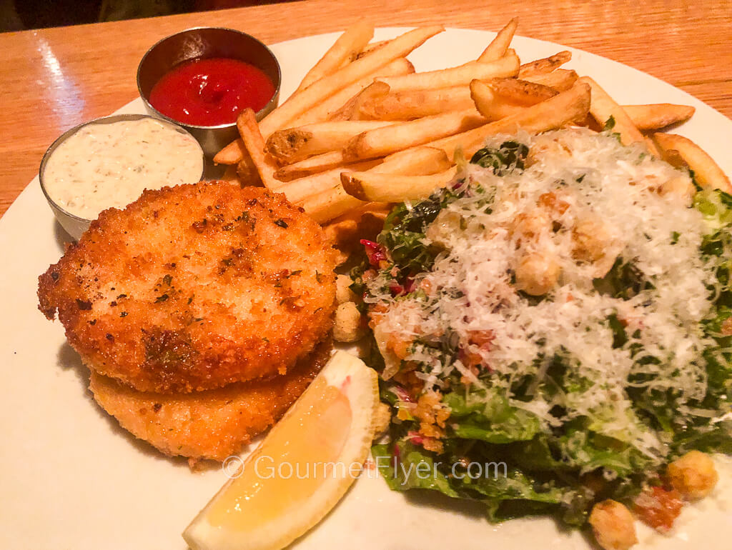 A stack of two fried crab cakes is served with French fries and a salad topped with shredded cheeses.