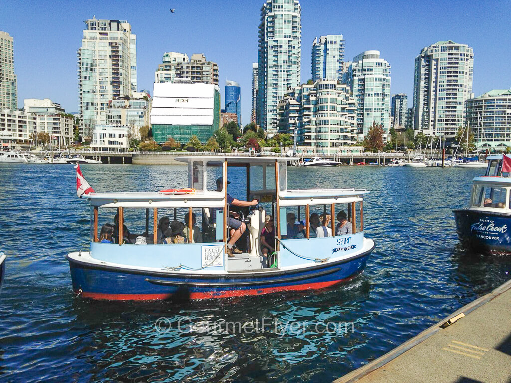 A small blue ferry full of passengers is off the docks in the water, with the skyline of Vancouver in the background.