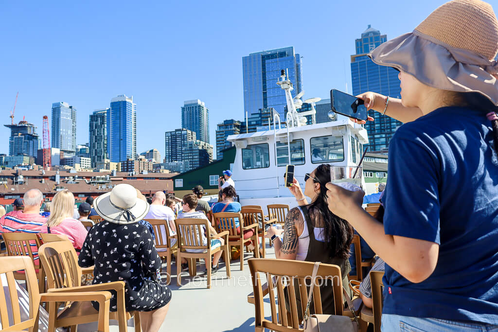 Engaging tourists on the sun deck of the boat are taking pictures with their cell phones, with Seattle's skyline in the background.