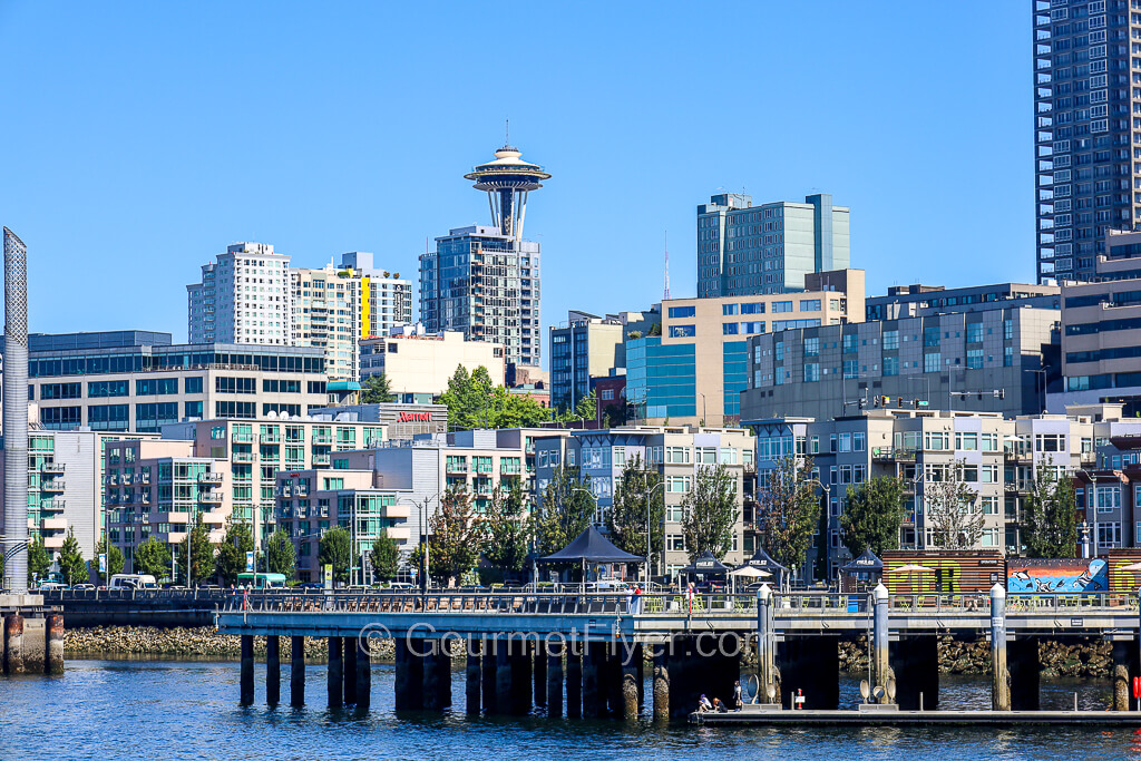 A community space is seen on a pier extending out to the water. The top of the Space Needle can be seen in the background.