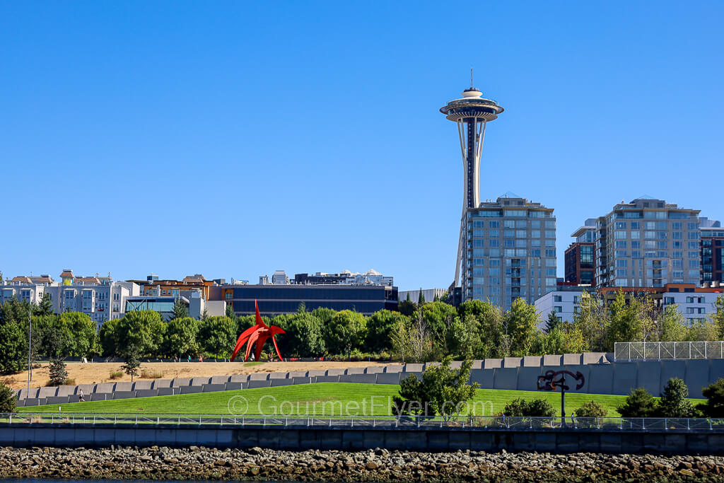 A park with plenty of green space has an iconic red sculpture sitting on a sandy area. The Space Needle can be seen clearly in the background.