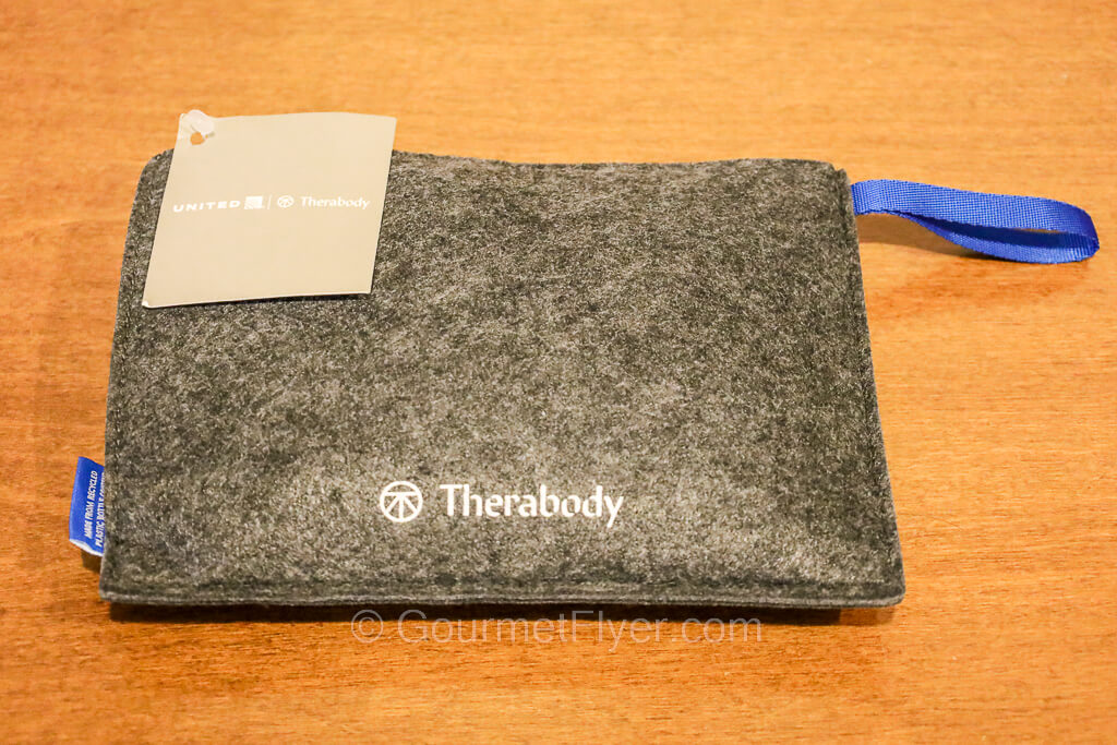 A gray travel pouch with the Therabody logo printed on the bottom and a United tag attached to it.