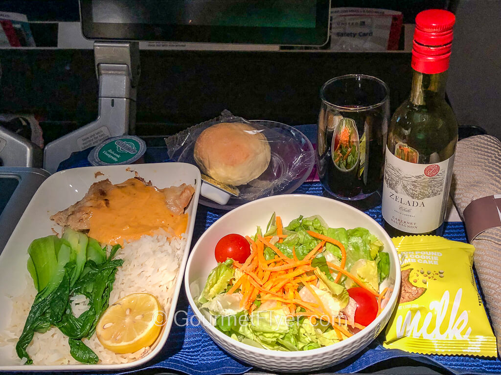 A dinner tray contains a plate of chicken served with gravy on rice, a green salad, and a mini bottle of red wine with a wine glass.
