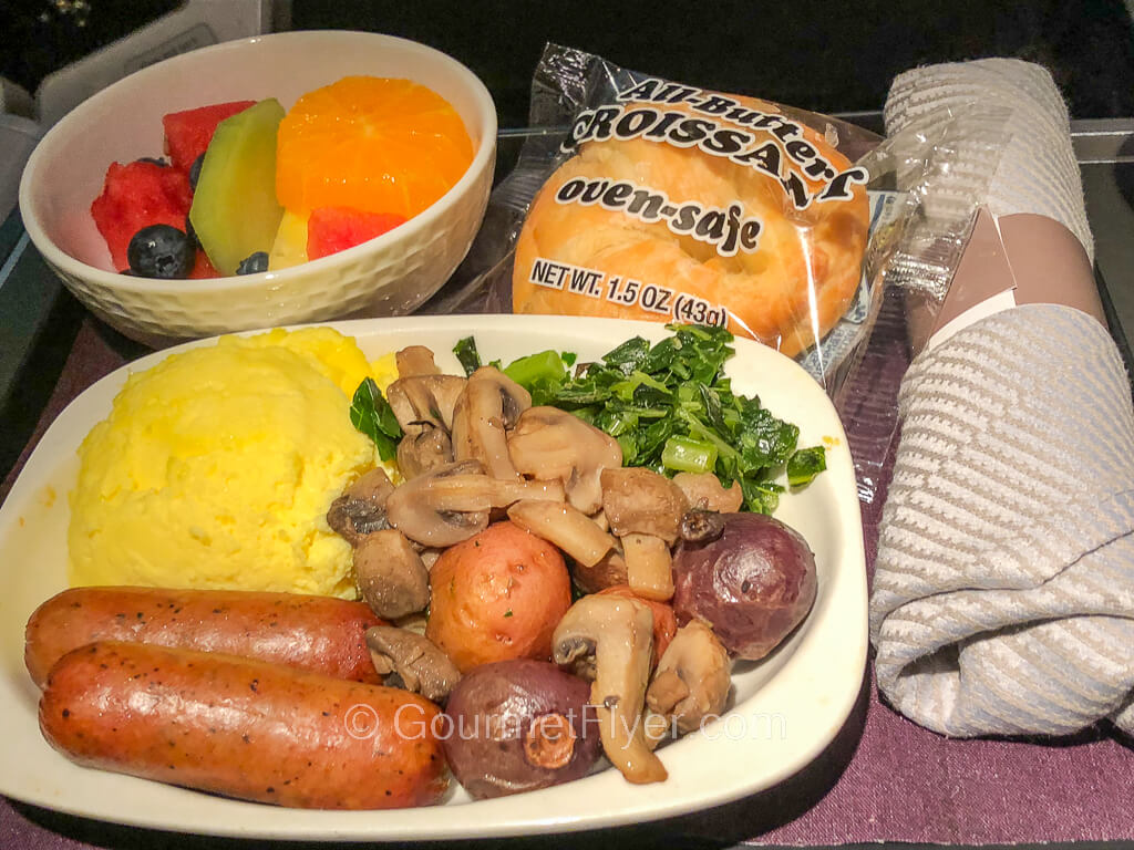 A plate of sausage and eggs served with mushrooms and potatoes is accompanied by a bowl of cut fruits. A rollup of utensils is on the right side of the tray.