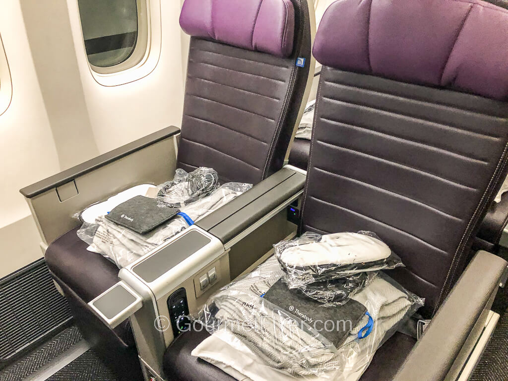 A pair of purple seats on the window side is stocked with a pile of pillows, blankets, slippers, a pouch for an amenity kit and a headset in plastic wraps.