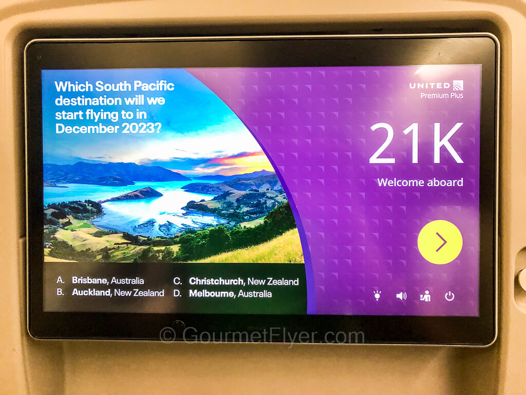 A seatback entertainment screen shows a South Pacific Island and the seat number 21K.