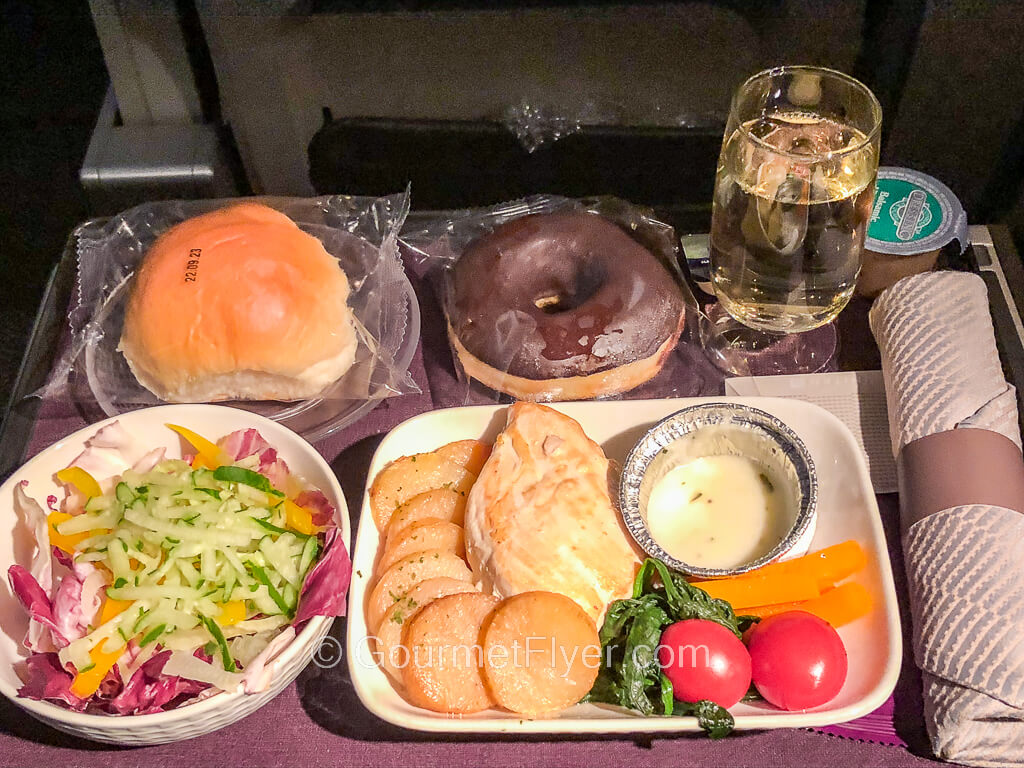 A dinner tray contains a plate of grill chicken served with sliced potatoes, a green salad, dinner roll, a chocolate donut, and a glass of white wine.