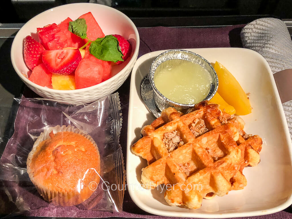 Golden brown waffles were served with apple sauce and accompanied by a bowl of fruits and a packaged pastry.