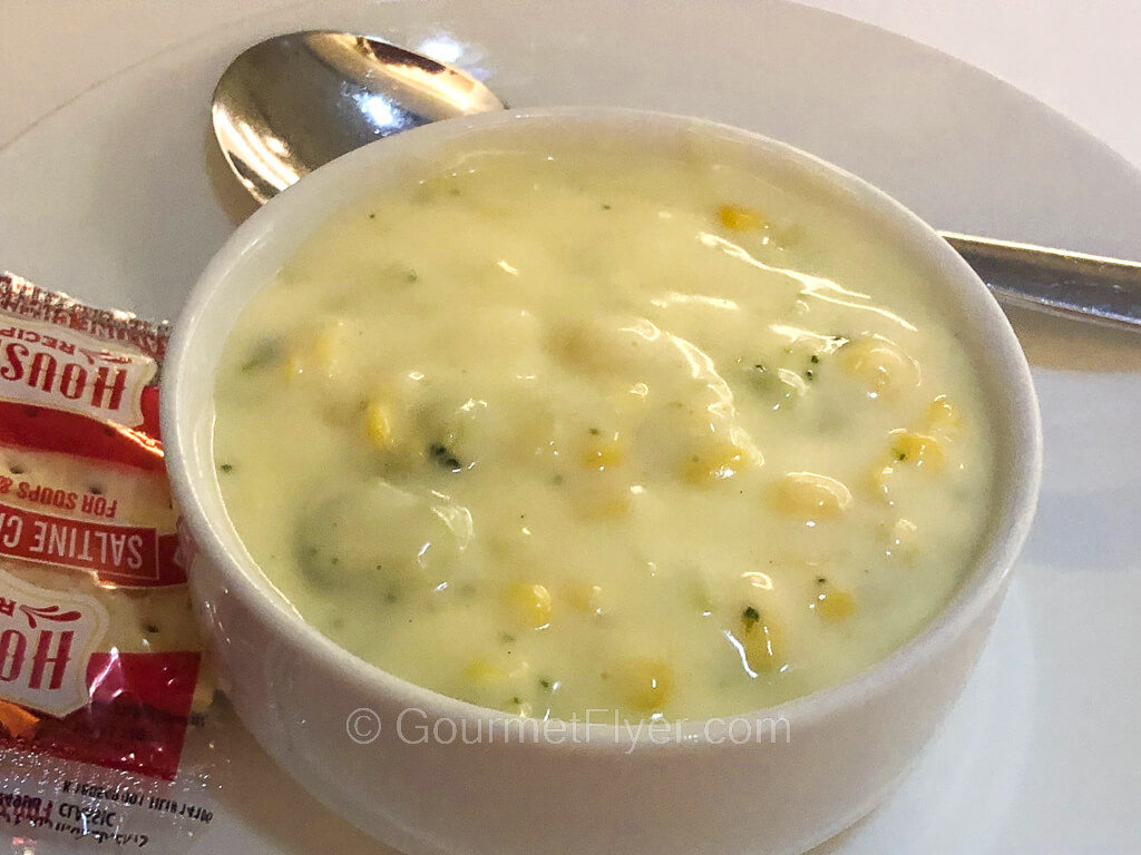 A bowl creamy and cheesy looking broccoli soup is placed on a small plate with a packet of crackers and a soup spoon.