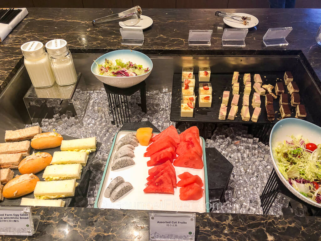 On top of a sunken area with ice are trays of finger sandwiches, sliced watermelons and dragon fruits and a bowl of salad in front, with a variety of cakes in the back.