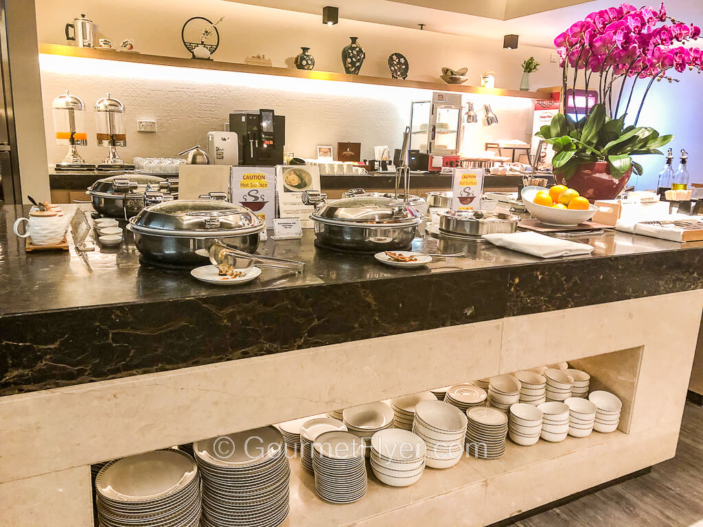 An extensive marble buffet counter has large chaffing dishes on one end is decorated with orchids on the other. Plates and bowls are stored below the counter.