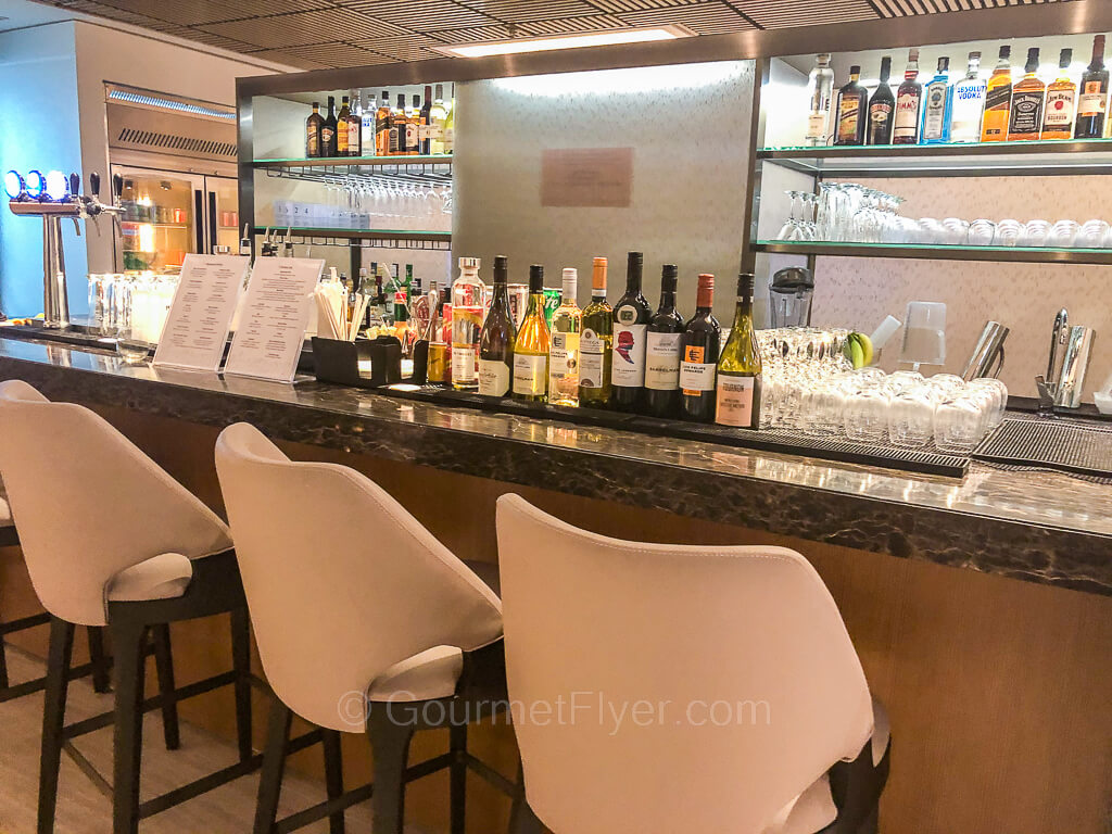 A full bar with displays of bottles of wines and liquors has a few barstools in front of it.