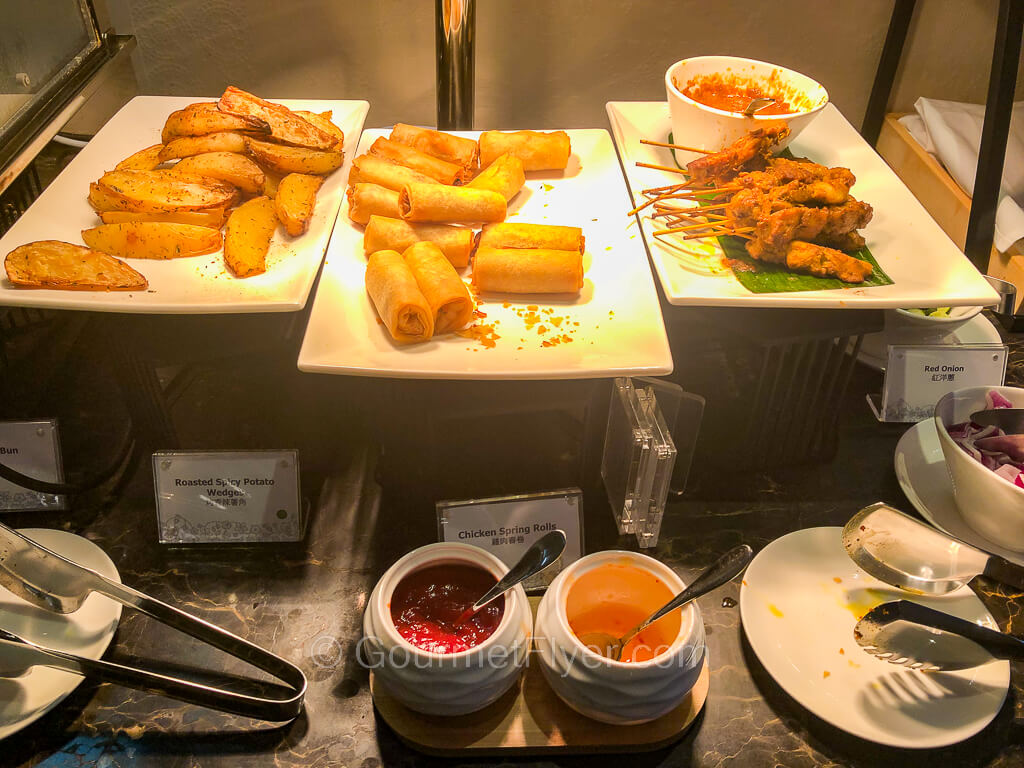 Three trays of food sit under heat lamps and are accompanied by sauces in containers with spoons.