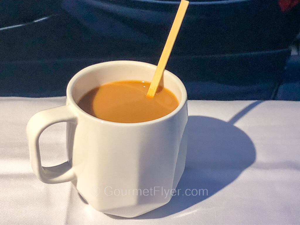 A white mug of coffee with cream and sugar sits atop the passenger's tray table.