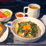 Review of Delta Airlines' first class features a meal tray with a quiche accompanied by fresh fruits, a croissant, and a mug of coffee.