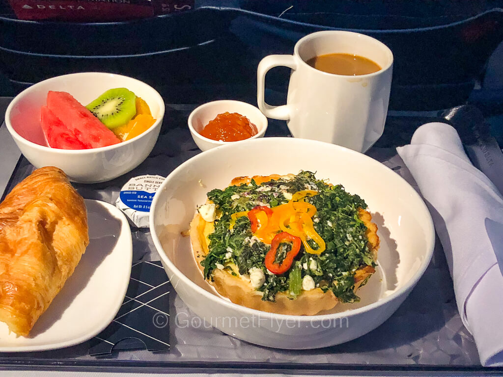Review of Delta Airlines' first class features a meal tray with a quiche accompanied by fresh fruits, a croissant, and a mug of coffee.