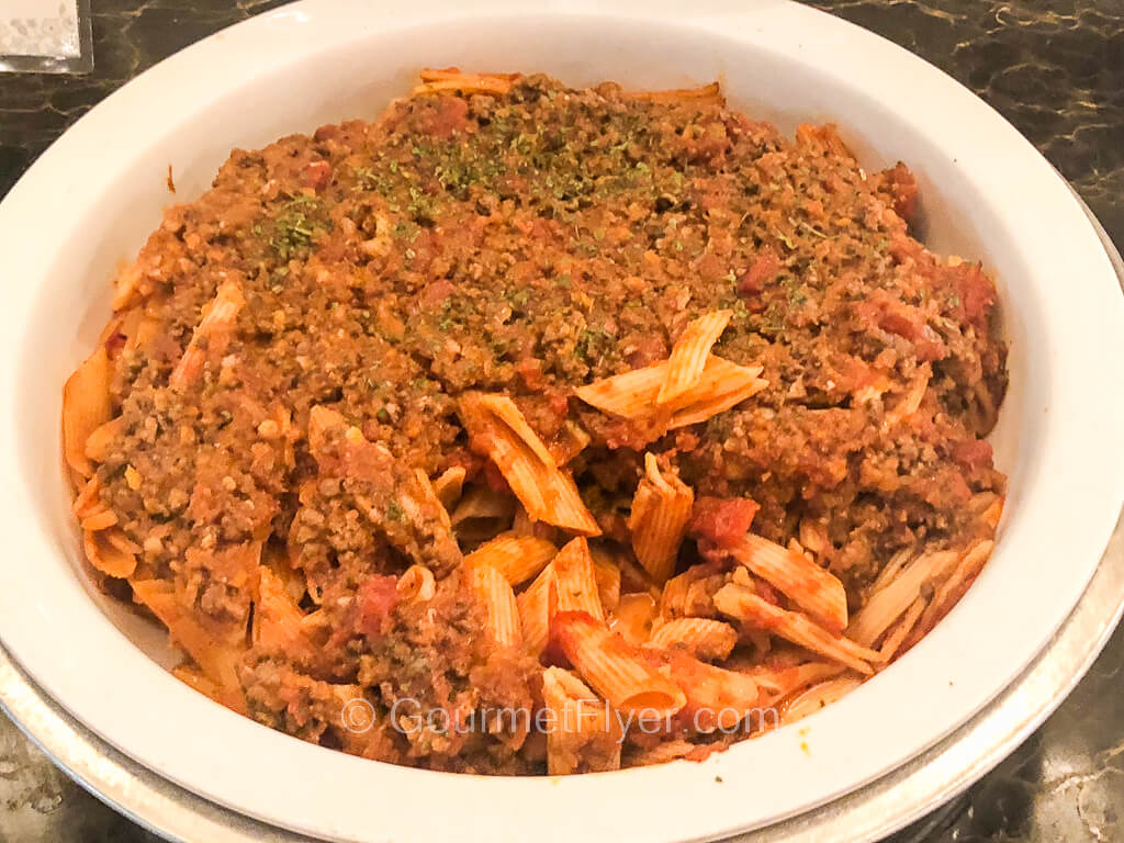 A round serving bowl is filled with penne pasta topped with a thick layer of meat and tomato sauce.