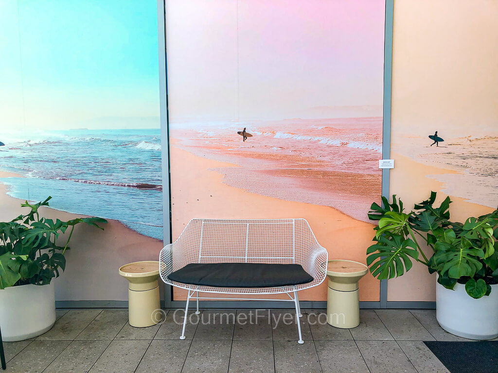 Three colorful panels of surfer-themed paintings are posted on the wall of the terrace, accompanied by green plants on their two sides and a chair in the middle.