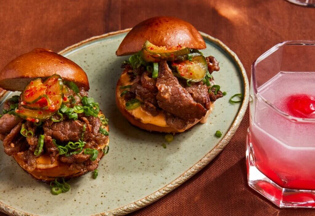 Two sliders with a tall stack of meat garnished with green onions sandwiched between a bun and served semi-open face.